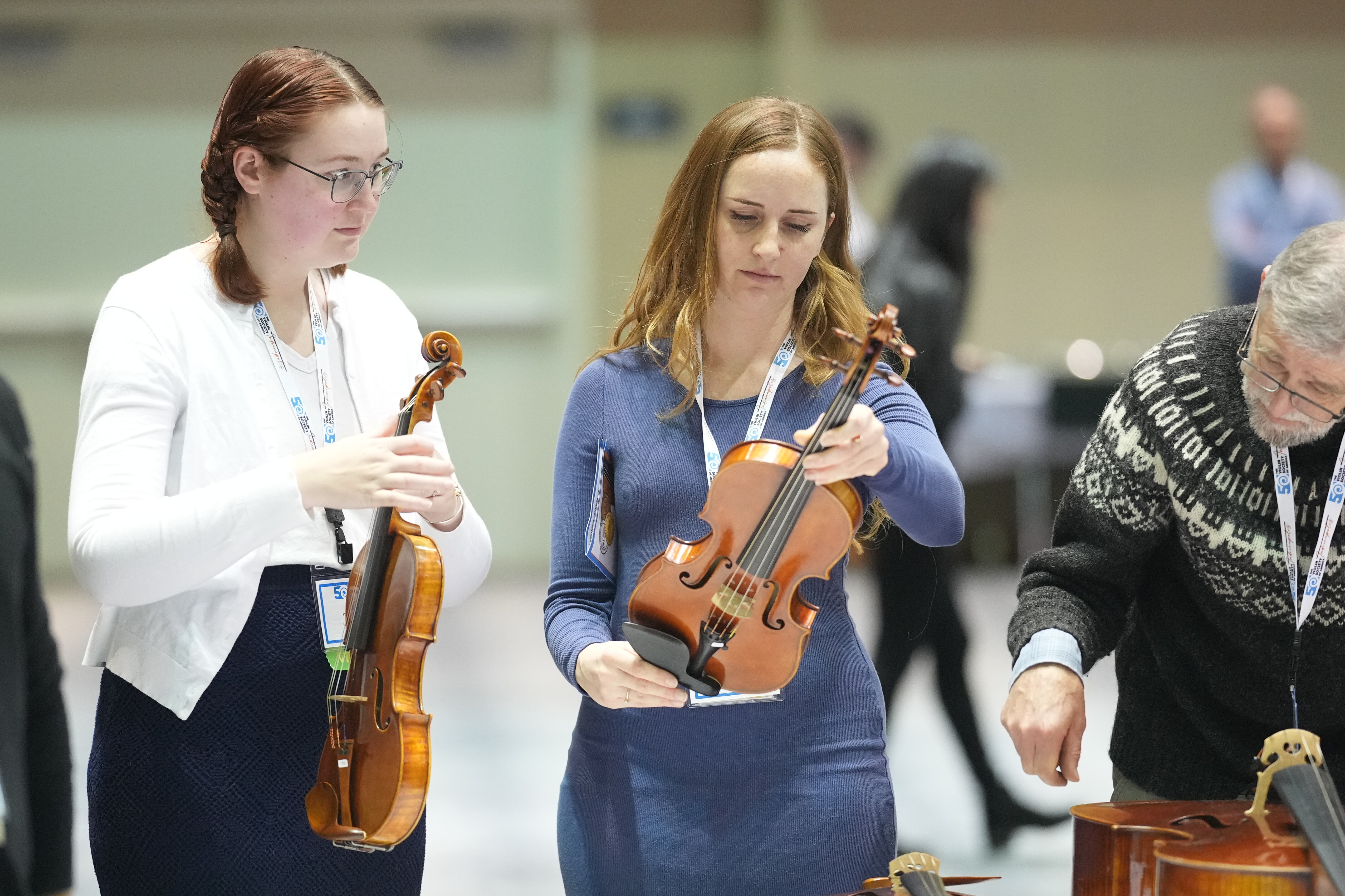 Two women examine violins at a music event, with a man nearby working on an instrument.