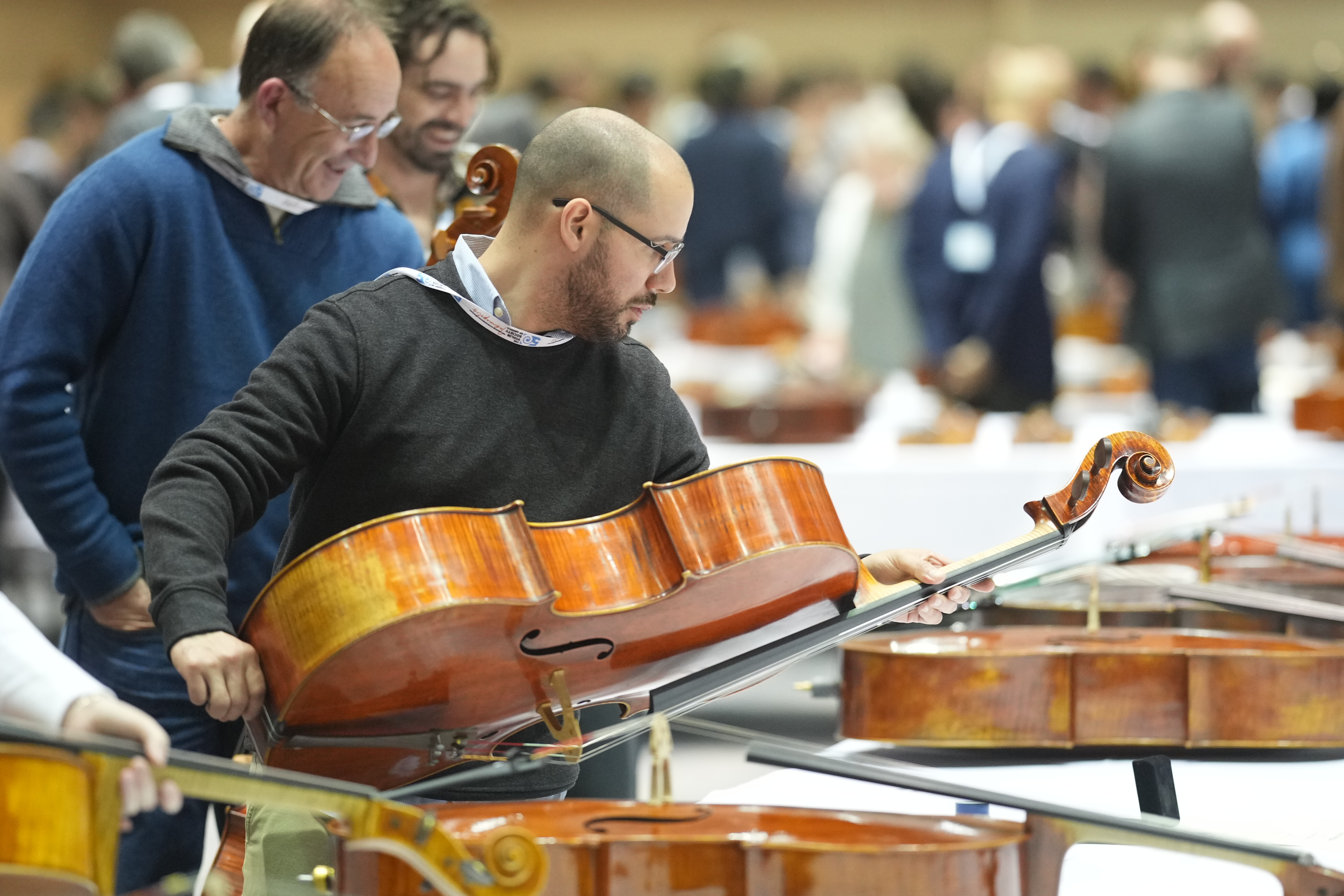 A man inspects a cello at a music event surrounded by others and various instruments.