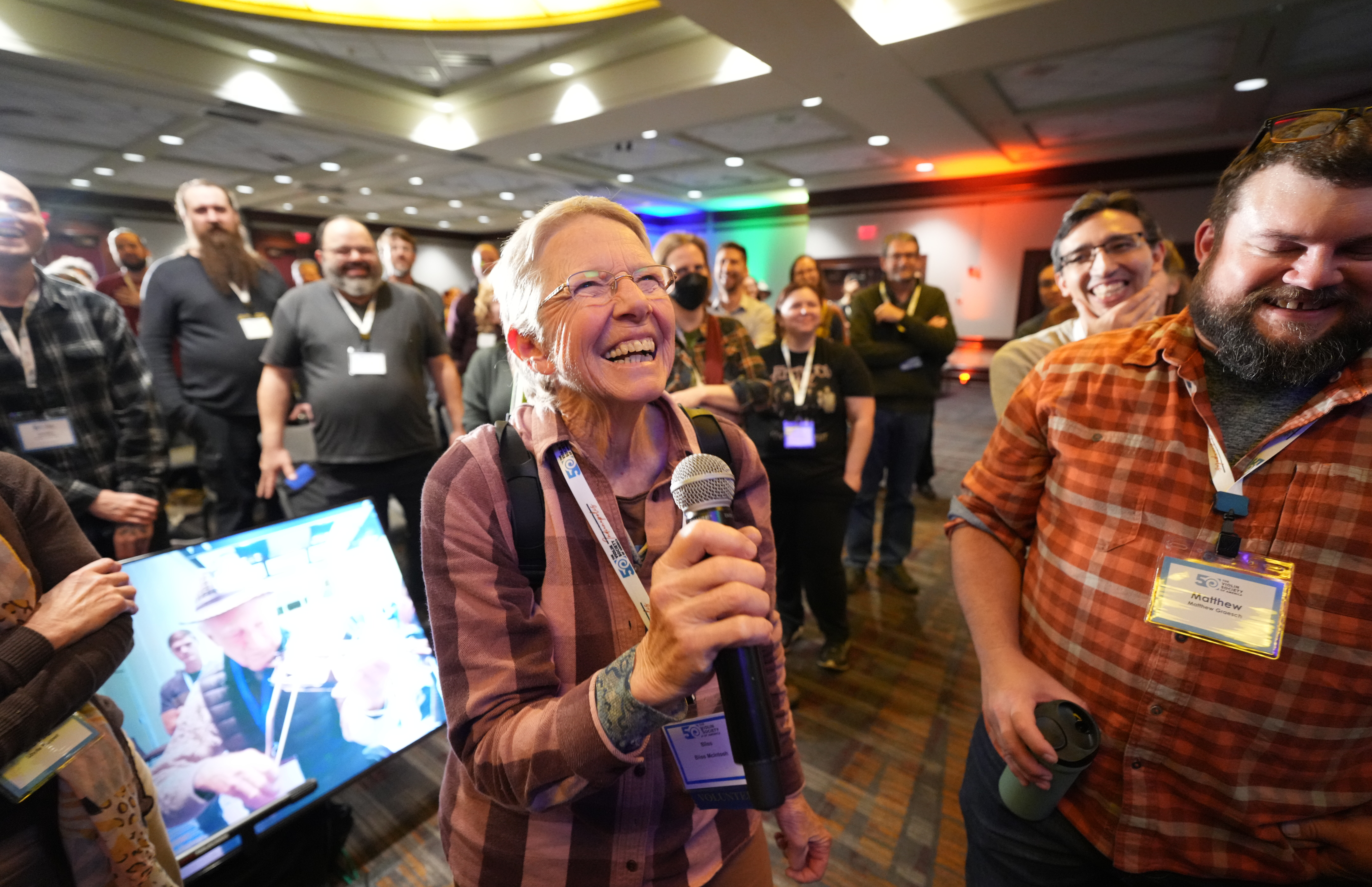 A smiling woman holds a microphone in a lively crowd at an event.