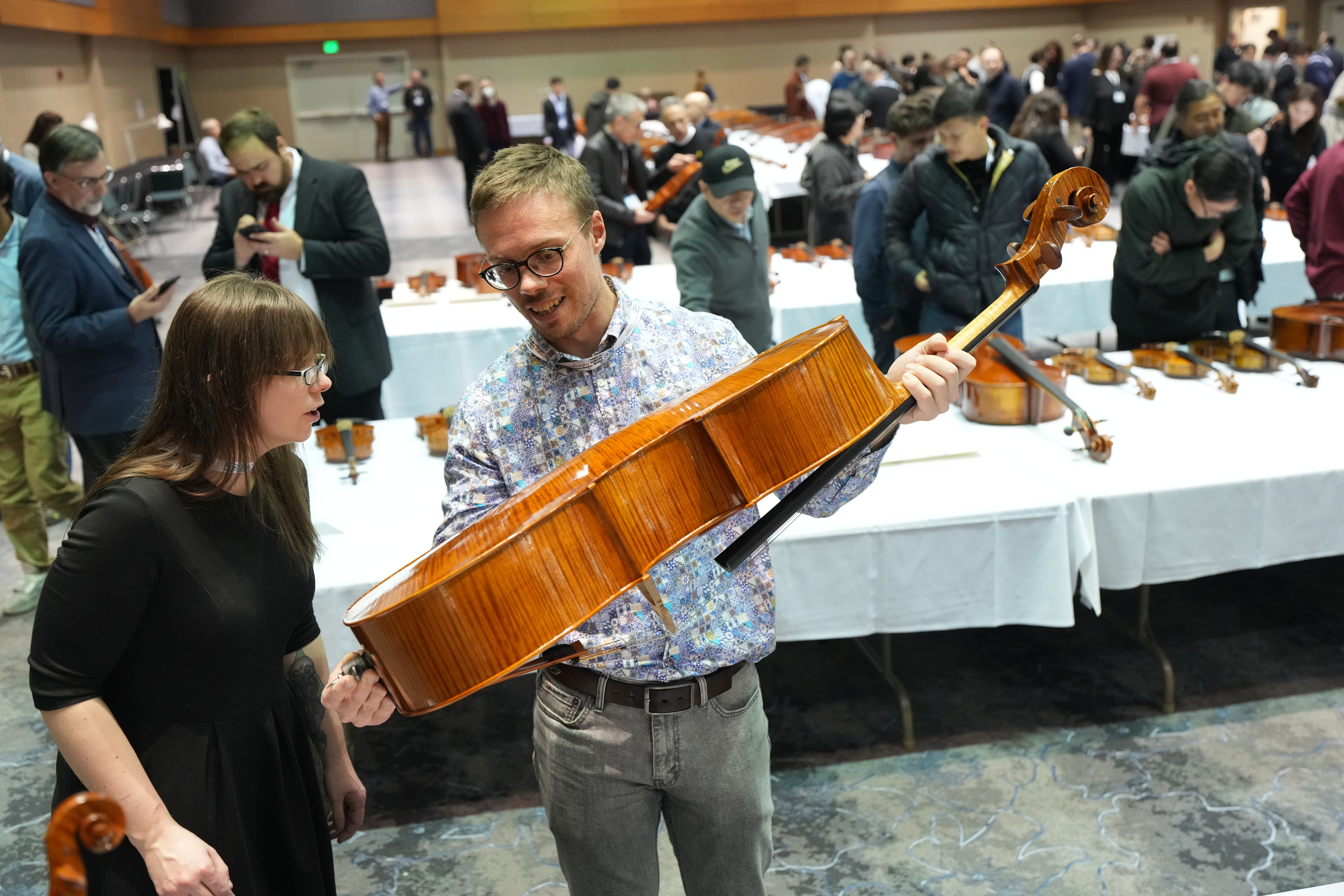 Two people examine a string instrument at a crowded event with various instruments displayed.