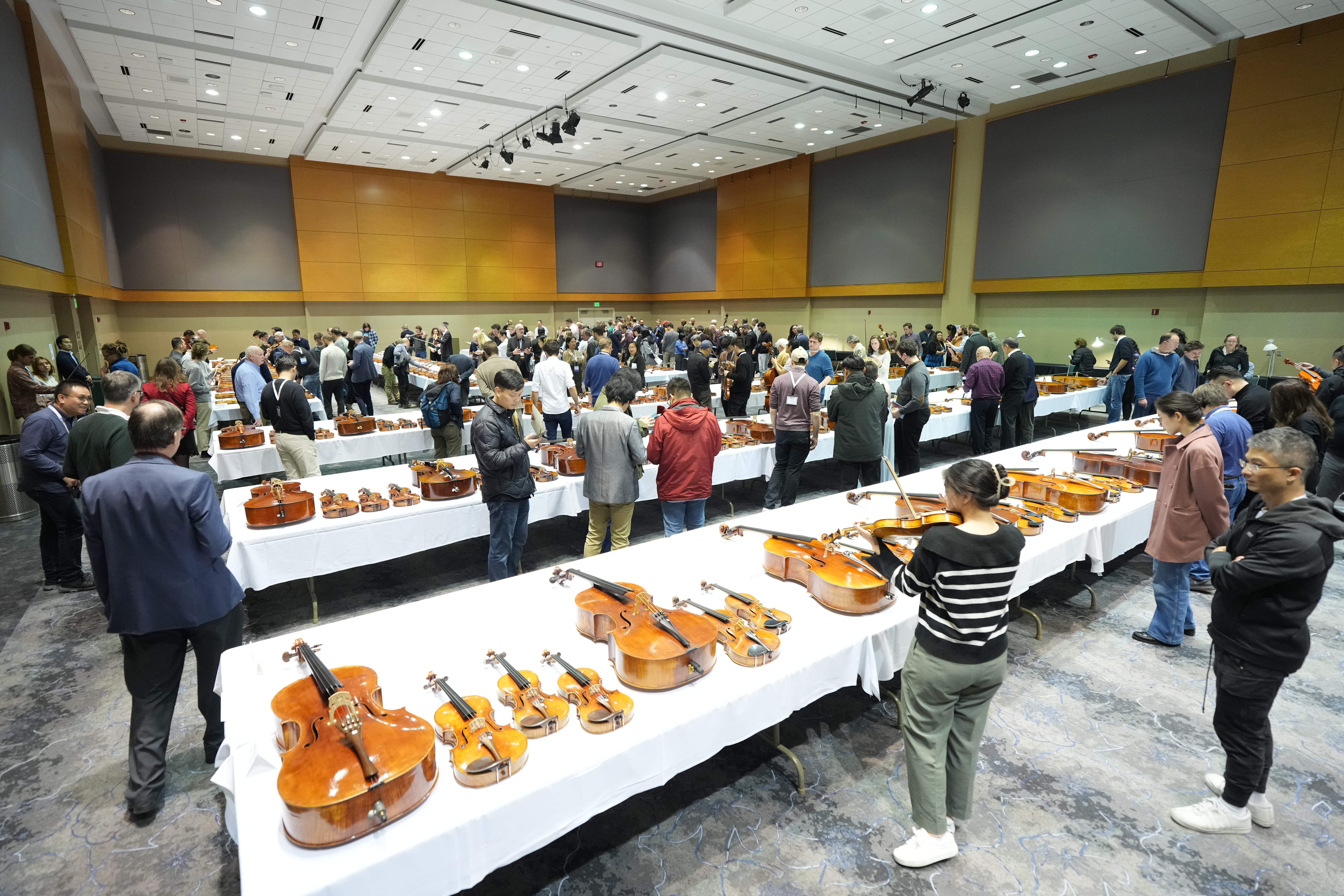 Crowd examining violins and cellos at a music event in a large venue.