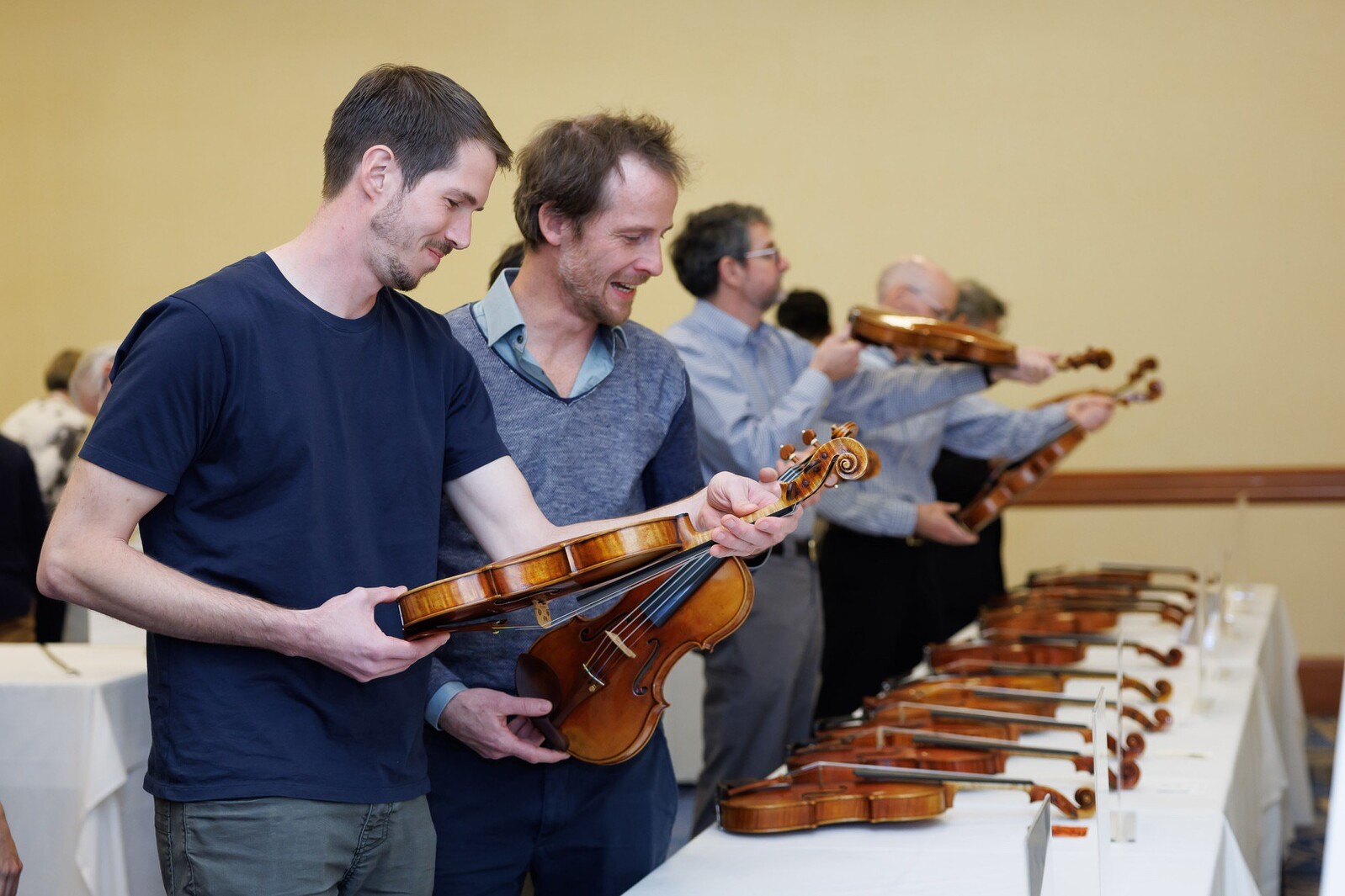 Two men examine violins at an event, with others in the background holding violins.