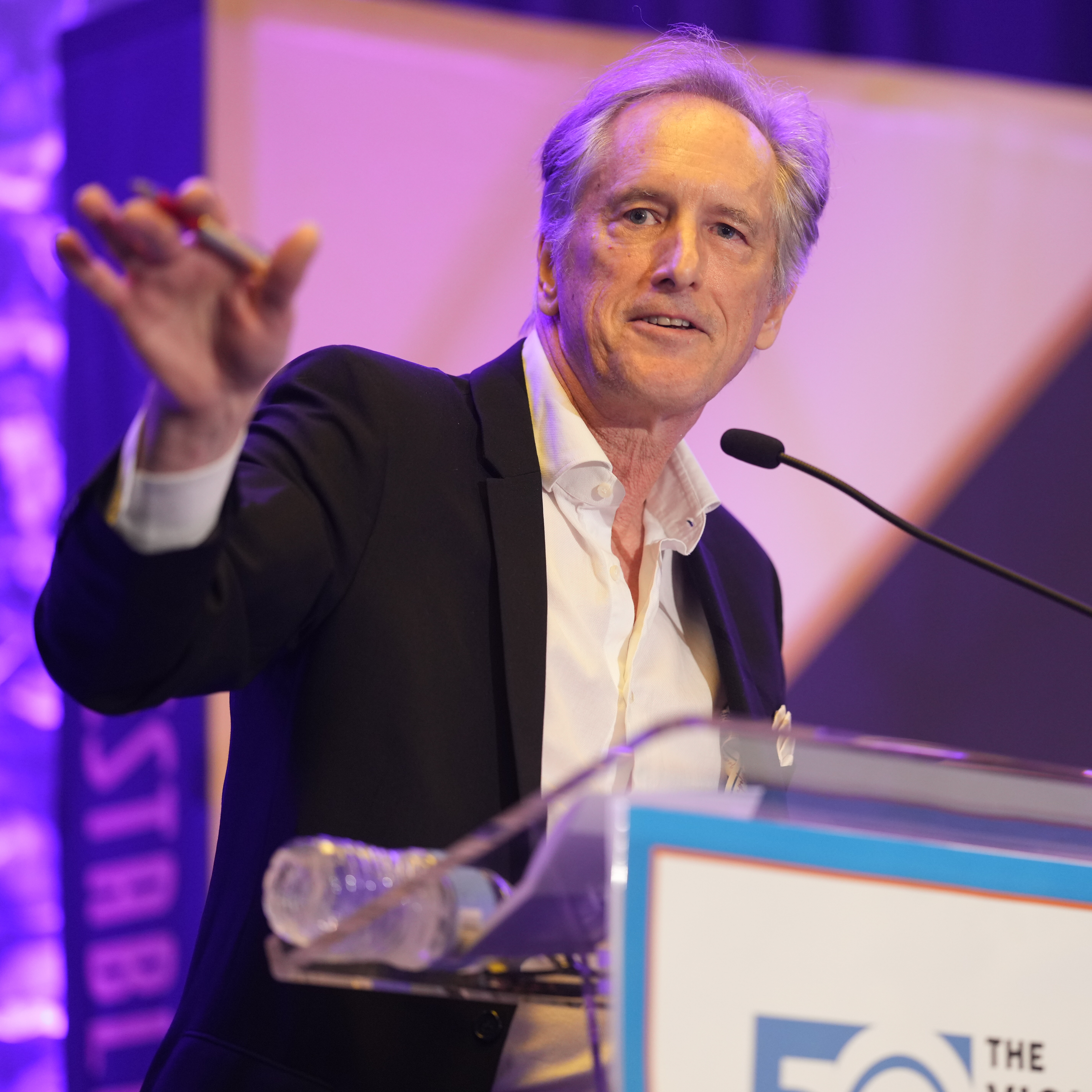 Man in a suit gestures while speaking at a podium during an event.